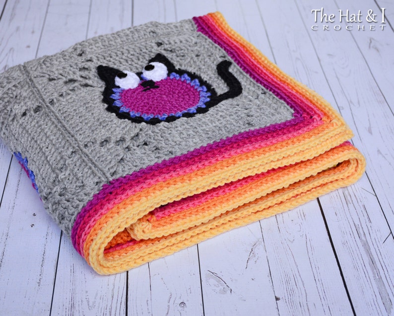 May include: A gray crochet baby blanket with a colorful border. The blanket features a crocheted cat applique with pink, purple, and blue details.