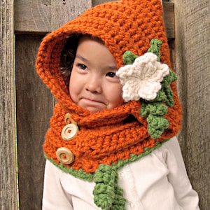 Può includere: Un set di cappello e sciarpa all'uncinetto in arancio e verde con un accento di fiore bianco. Il cappello ha un cappuccio e la sciarpa ha bottoni di legno.