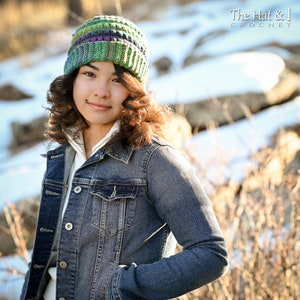 May include: A person wearing a green, purple, and white striped crochet beanie hat. The hat is made with a textured stitch and has a slouchy fit. The person is wearing a denim jacket and has brown hair.