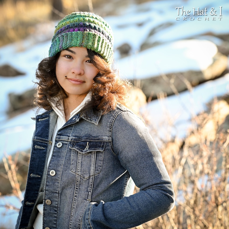 May include: A person wearing a green, purple, and white striped crochet beanie hat. The hat is made with a textured stitch and has a slouchy fit. The person is wearing a denim jacket and has brown hair.