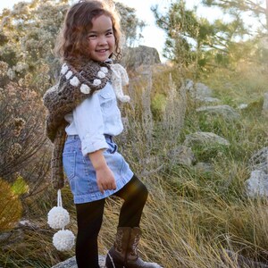 May include: A young person wearing a brown and white crocheted shawl with pom-poms, a white button-down shirt, a blue denim skirt, black tights, and brown cowboy boots. The person is standing on a rock in a grassy field.