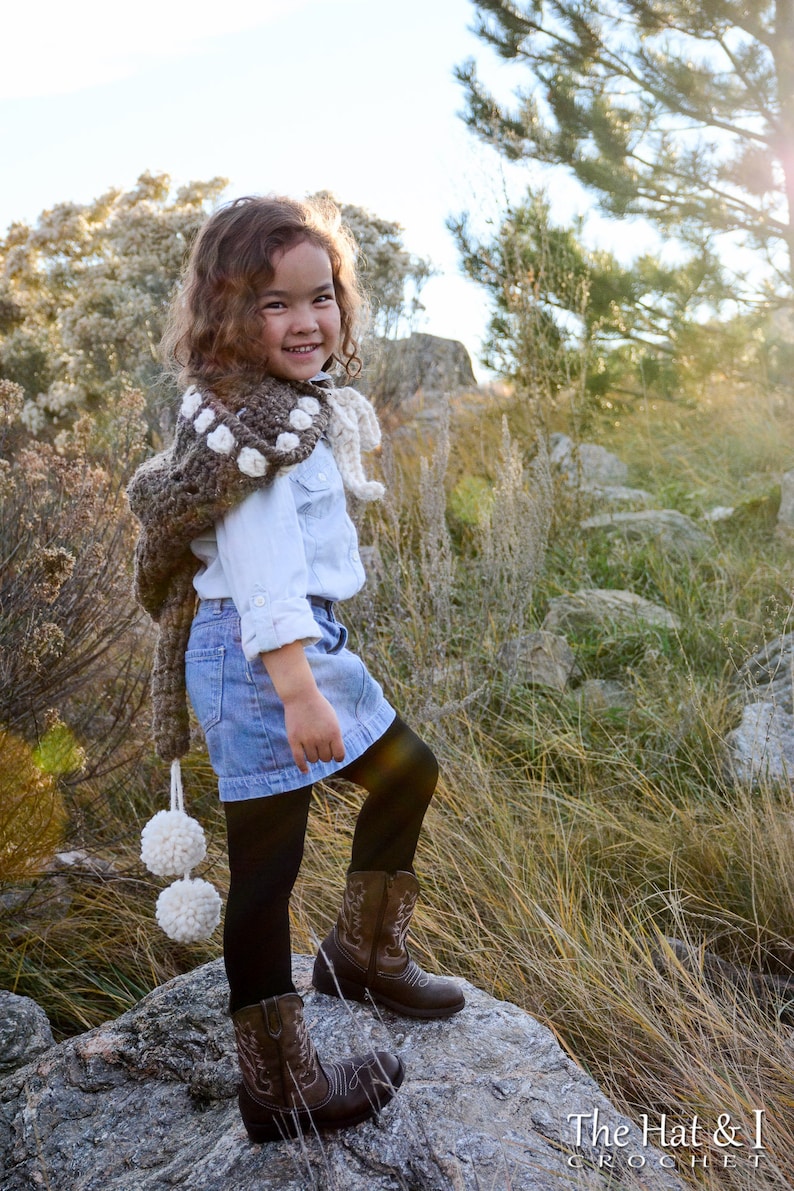May include: A young person wearing a brown and white crocheted shawl with pom-poms, a white button-down shirt, a blue denim skirt, black tights, and brown cowboy boots. The person is standing on a rock in a grassy field.