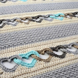 May include: A close-up of a crocheted blanket with a repeating pattern of hearts in shades of gray, beige, and turquoise. The blanket is made with a textured stitch and has a soft, cozy look.