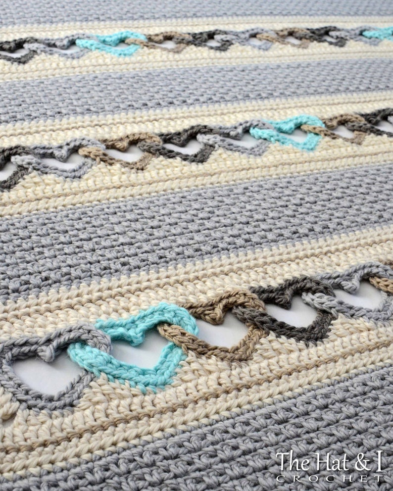 May include: A close-up of a crocheted blanket with a repeating pattern of hearts in shades of gray, beige, and turquoise. The blanket is made with a textured stitch and has a soft, cozy look.