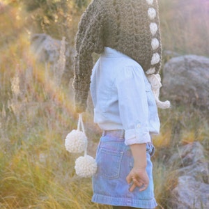 May include: A brown crocheted hooded hat with white pom-poms and white crocheted accents. The hat is worn by a child in a blue shirt and denim skirt.