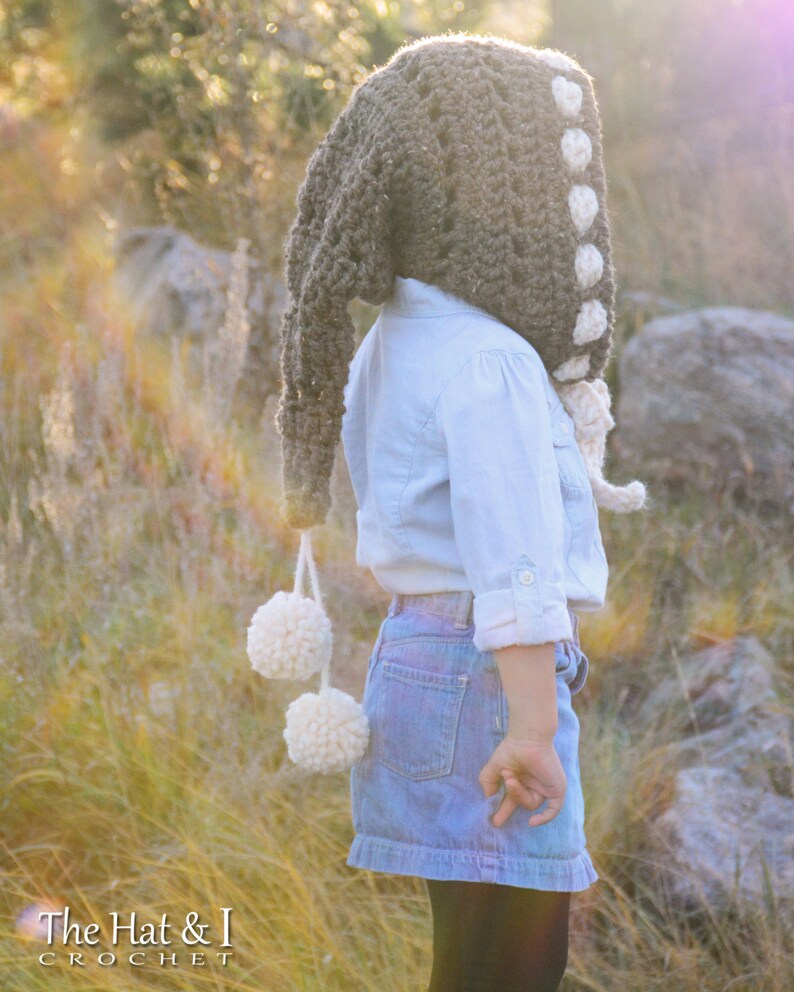 May include: A brown crocheted hooded hat with white pom-poms and white crocheted accents. The hat is worn by a child in a blue shirt and denim skirt.