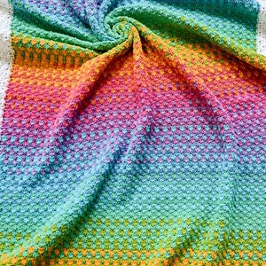 May include: A rainbow ombre crochet baby blanket with a white border. The blanket is made with a textured stitch and is folded in a spiral shape.