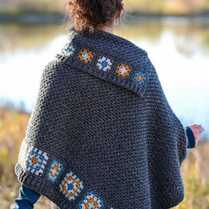 May include: A dark gray crochet poncho with colorful floral accents. The poncho features a square pattern with blue, orange, and white flowers along the edges and collar. The Hat & I Crochet logo is visible in the upper right corner.