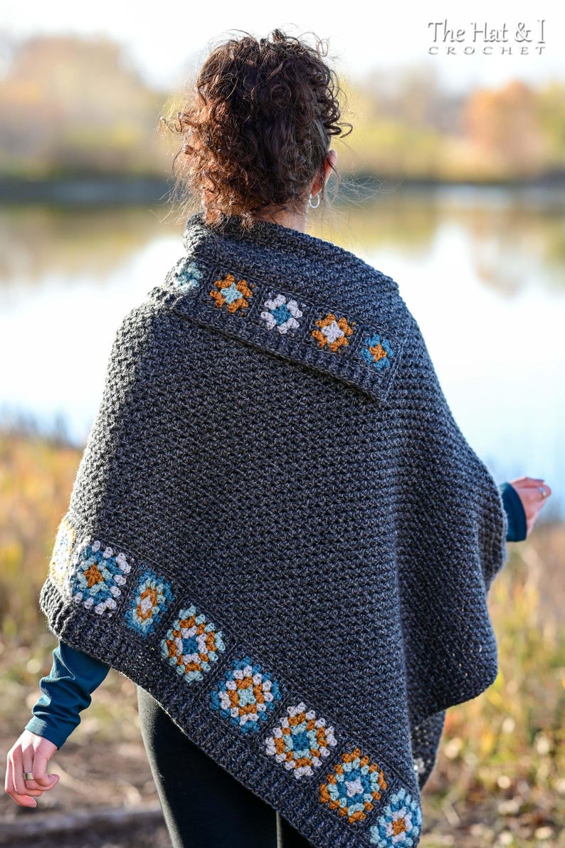 May include: A dark gray crochet poncho with colorful floral accents. The poncho features a square pattern with blue, orange, and white flowers along the edges and collar. The Hat & I Crochet logo is visible in the upper right corner.