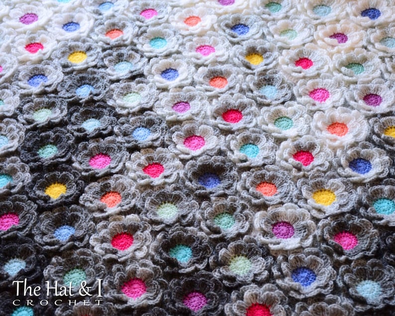 Puede incluir: Una manta de ganchillo con un degradado de gris a blanco, que presenta un patr&oacute;n repetitivo de flores de colores. Las flores est&aacute;n tejidas a ganchillo en varios tonos de rosa, azul, verde, amarillo y naranja.