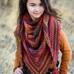 Puede incluir: Una mujer lleva una chalina de ganchillo de colores con un detalle de flecos. La chalina est&aacute; hecha con una variedad de tonos de rojo, naranja, amarillo, verde y morado. La mujer lleva una camisa naranja de manga larga y tiene el pelo largo y oscuro.