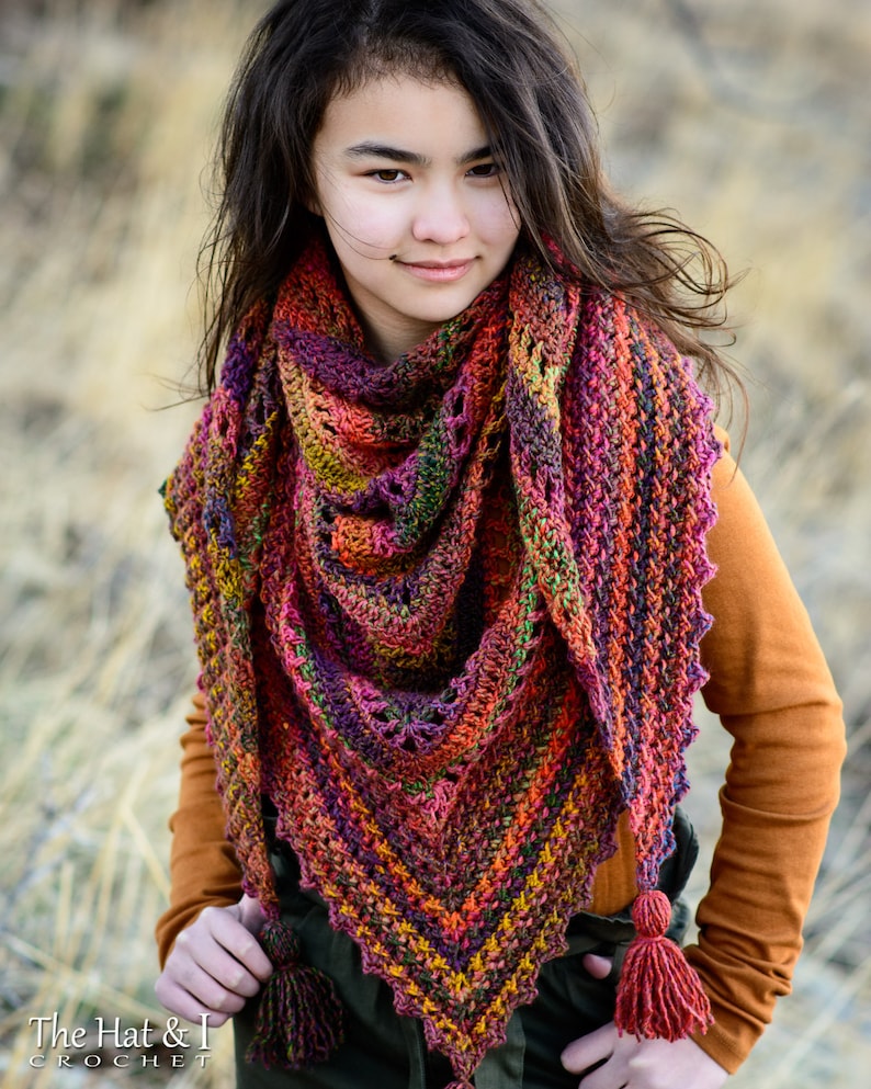 Puede incluir: Una mujer lleva una chalina de ganchillo de colores con un detalle de flecos. La chalina est&aacute; hecha con una variedad de tonos de rojo, naranja, amarillo, verde y morado. La mujer lleva una camisa naranja de manga larga y tiene el pelo largo y oscuro.
