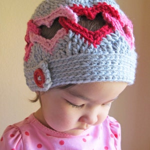 Puede incluir: Un gorro de punto gris con corazones rosas y rojos. El gorro tiene un botón en el lateral.