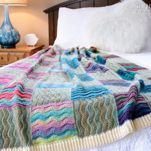 Pode incluir: Uma manta de croch&ecirc; colorida com um padr&atilde;o ondulado em tons de azul, verde, rosa e roxo. A manta tem uma borda de croch&ecirc; branca e est&aacute; drapeada sobre uma cama branca.