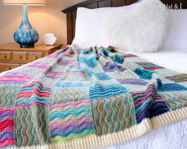 Pode incluir: Uma manta de croch&ecirc; colorida com um padr&atilde;o ondulado em tons de azul, verde, rosa e roxo. A manta tem uma borda de croch&ecirc; branca e est&aacute; drapeada sobre uma cama branca.