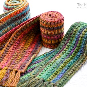 Puede incluir: Tres bufandas de ganchillo en tonos de verde, naranja y marr&oacute;n con un punto de relieve de pomp&oacute;n. Las bufandas est&aacute;n enrolladas y tienen borlas en los extremos. Las bufandas est&aacute;n hechas por The Hat & I Crochet.