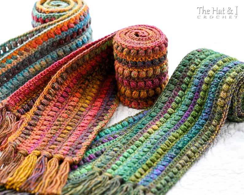 Puede incluir: Tres bufandas de ganchillo en tonos de verde, naranja y marr&oacute;n con un punto de relieve de pomp&oacute;n. Las bufandas est&aacute;n enrolladas y tienen borlas en los extremos. Las bufandas est&aacute;n hechas por The Hat & I Crochet.