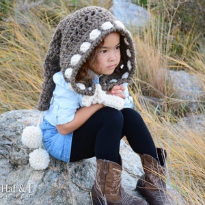 May include: A young girl wearing a brown and white crocheted hooded scarf with pom-poms. The scarf has a white polka dot pattern.