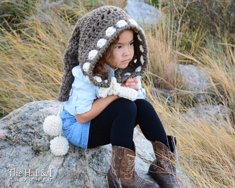 May include: A young girl wearing a brown and white crocheted hooded scarf with pom-poms. The scarf has a white polka dot pattern.