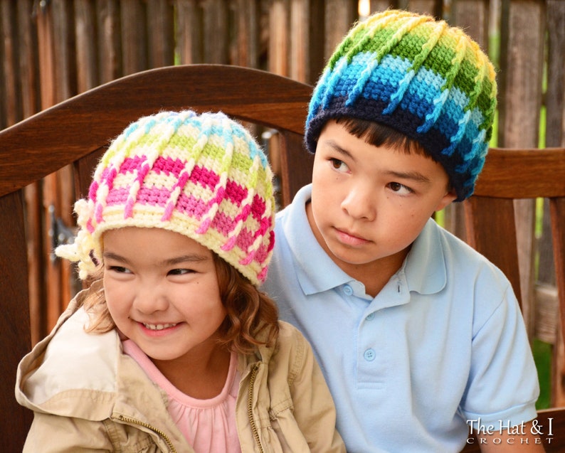 Pu&ograve; includere: Due bambini indossano cappelli all'uncinetto colorati. La ragazza indossa un cappello rosa, bianco e verde con un dettaglio floreale. Il ragazzo indossa un cappello blu, verde e giallo. I bambini sono seduti su una panca di legno.