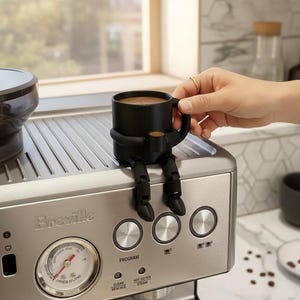 May include: A black coffee mug with attached arms and legs, sitting on a silver espresso machine. The mug is filled with coffee. The espresso machine has a pressure gauge and control buttons.