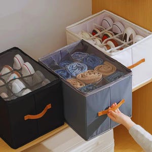 May include: Three fabric storage boxes with lids, organized on a shelf. One black box contains shoes, a gray box holds rolled clothing, and a white box also has shoes. Each box has a brown handle.