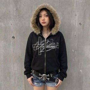 May include: Black zip-up hoodie with a faux fur-lined hood. The hoodie features a silver cross and script design on the front. A studded belt with a skull buckle is worn with denim shorts. The hoodie has a relaxed fit.