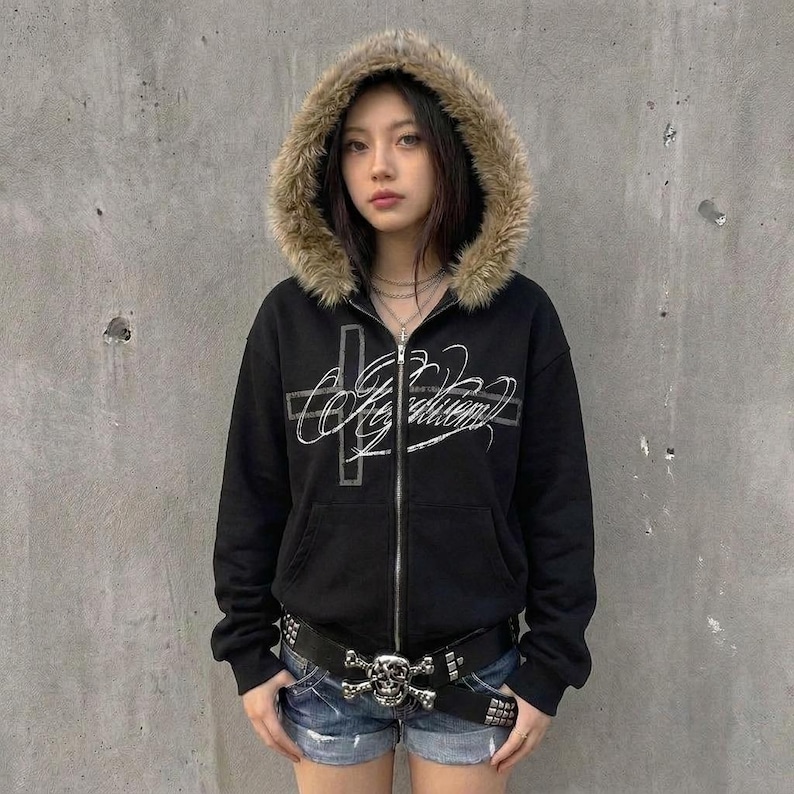 May include: Black zip-up hoodie with a faux fur-lined hood. The hoodie features a silver cross and script design on the front. A studded belt with a skull buckle is worn with denim shorts. The hoodie has a relaxed fit.
