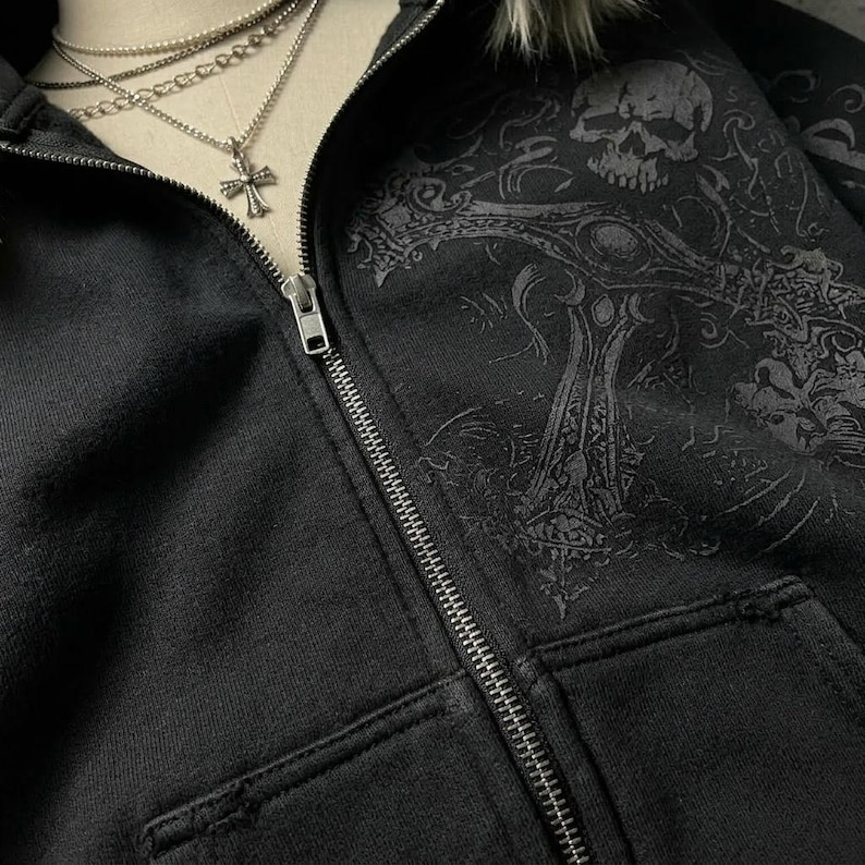 Op de afbeelding: Zwarte hoodie met ritssluiting en een distressed design. De hoodie heeft een schedel- en kruisafbeelding op de voorkant. Een zilveren kruis hanger hangt aan een gelaagde ketting.