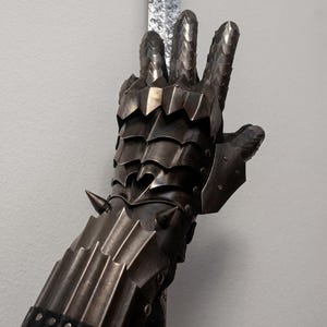 Peut inclure: Gros plan d'un gant de métal sombre et détaillé avec des doigts articulés et un poignet à pointes. L'armure présente un design en couches avec un aspect texturé et vieilli. Une partie d'une lame texturée est visible en arrière-plan.