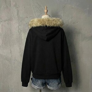 May include: A black hooded sweatshirt with a faux fur trim around the hood. The sweatshirt is displayed on a mannequin and paired with denim shorts. The background is a plain gray wall.