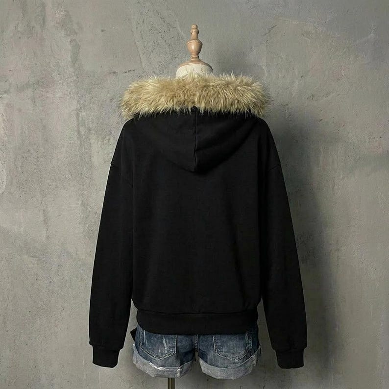 May include: A black hooded sweatshirt with a faux fur trim around the hood. The sweatshirt is displayed on a mannequin and paired with denim shorts. The background is a plain gray wall.