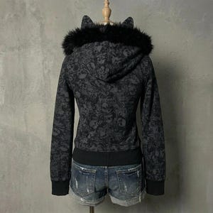 K&ouml;nnte beinhalten: Ein schwarzer Kapuzenpullover mit Rei&szlig;verschluss, Totenkopfmuster, Kunstfellbesatz und Katzenohren an der Kapuze. Der Kapuzenpullover hat einen schwarzen gerippten Saum und B&uuml;ndchen. Das Model tr&auml;gt Jeansshorts.