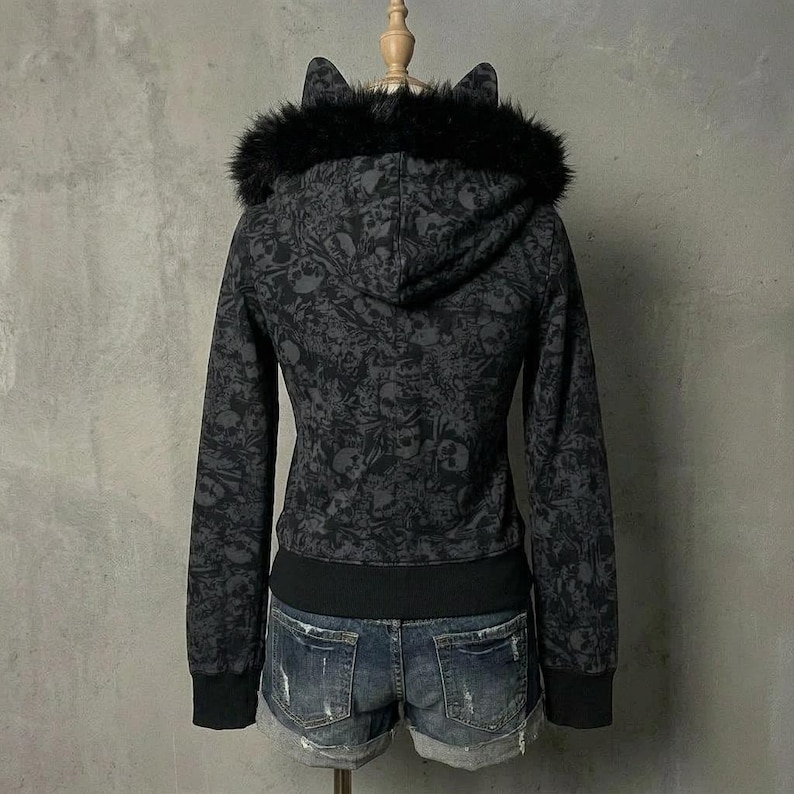 K&ouml;nnte beinhalten: Ein schwarzer Kapuzenpullover mit Rei&szlig;verschluss, Totenkopfmuster, Kunstfellbesatz und Katzenohren an der Kapuze. Der Kapuzenpullover hat einen schwarzen gerippten Saum und B&uuml;ndchen. Das Model tr&auml;gt Jeansshorts.