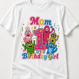 May include: A white t-shirt featuring a colorful graphic with the text "Mom of the 1st Birthday Girl". The design includes a large pink glittery number one surrounded by cartoon characters including a pink creature, a green striped monster, a yellow robot, and a blue cat-like figure, all accented with confetti.