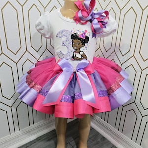 Op de afbeelding: Een wit shirt met korte mouwen voorzien van een groot glitterend cijfer 3 en een stripfiguur met een feesthoedje, gecombineerd met een meerlaagse tule rok in roze en paars met satijnen lintafwerking en een grote bijpassende haarstrik.