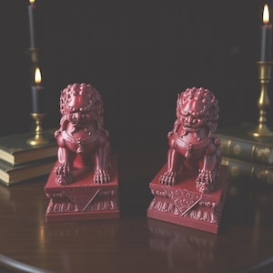 May include: Two red Foo Dog statues with intricate detailing, presented on ornate bases. They are placed on a wooden surface, with candles and books in the background, creating a vintage aesthetic.