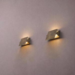 Op de afbeelding: Twee wandkandelaars met een geometrisch ontwerp. Elke kandelaar heeft een vierkante, metalen kap die naar beneden is gericht en een warm, geel licht uitstraalt. De wandkandelaars zijn gemonteerd op een gedempte paarse muur.