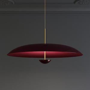 Op de afbeelding: Een moderne hanglamp met een bordeauxrode kap en een goudkleurige steel. De kap heeft een brede, ondiepe schijfvorm met een contrasterend rood interieur. De armatuur hangt aan een goudkleurige staaf.