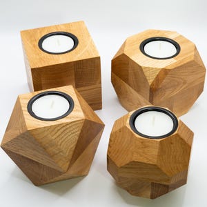 Può includere: Quattro portacandele in legno di varie forme geometriche. Ogni portacandela è realizzato in legno chiaro e presenta un inserto in metallo nero per contenere una candela bianca. Le forme includono un cubo, una sfera sfaccettata e altri due design angolari.