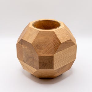 Può includere: Un vaso geometrico in legno con finitura naturale. Il vaso è costruito con più pannelli di legno angolati, creando una forma sferica sfaccettata. L'interno è cavo e l'apertura superiore è circolare.