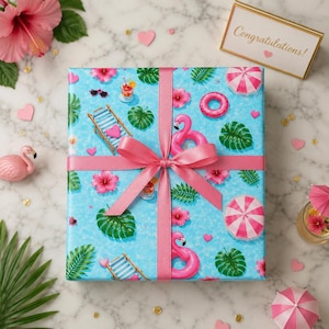 Puede incluir: Un regalo envuelto en papel azul con un estampado tropical de flamencos rosas, flores de hibisco y sombrillas de playa. El regalo está atado con una cinta y un lazo rosas. Una tarjeta dice "¡Enhorabuena!"