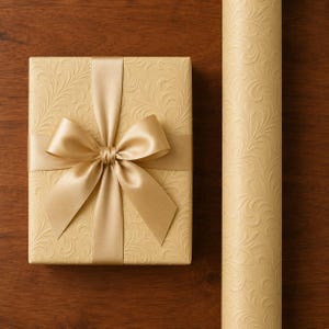 May include: A rectangular gift box wrapped in light gold paper with an embossed swirling leaf pattern, tied with a matching satin ribbon bow. Beside the box lies a matching roll of the same embossed light gold gift wrapping paper.