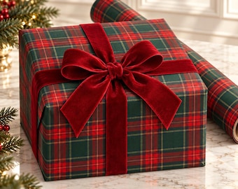 May include: A square gift box wrapped in dark green and red plaid paper, tied with a wide, deep red velvet ribbon bow. A matching roll of plaid wrapping paper sits behind the box, which is positioned on a white marble surface.