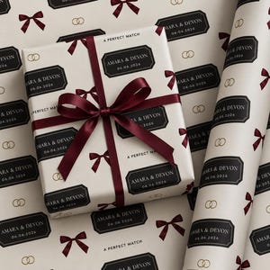Könnte beinhalten: Ein Geschenkpaket und eine Rolle Geschenkpapier mit einem wiederkehrenden Muster aus dunkelroten Schleifen, goldenen Eheringen und schwarzen rechteckigen Etiketten mit dem weißen Text AMARA & DEVON 06.06.2026 und A PERFECT MATCH. Das Paket ist mit einem Band.