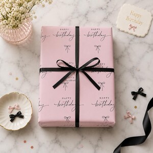 Puede incluir: Un regalo envuelto en papel rosa con la impresión "Happy Birthday" en escritura negra e ilustraciones de lazo. El regalo está atado con una cinta de raso negra. También hay una pequeña tarjeta con "Happy Birthday".
