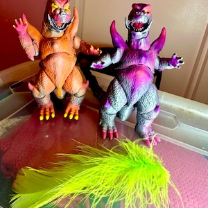 May include: Two colorful monster action figures with open mouths and outstretched arms. One is orange and yellow, the other is purple and gray. A bright green feather lies in front of them. The figures have sharp teeth and claws.