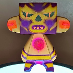 May include: A colorful, abstract wooden figure with a square head and a triangular body. The face is painted with a purple mask design, yellow accents, and orange highlights. The body is cream-colored with yellow and purple stripes, and a pink spray-painted design.