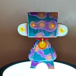 May include: A vibrant art toy with a square head and rounded body. The toy features a blue, purple, and orange colour scheme with a U-shaped design on the head. It has large, eye-shaped side pieces and a circular tag around its neck.