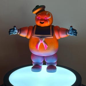 May include: A colorful, stylized figurine of a character with an orange body, blue hands and feet, and a black sailor collar. The figure has a smiling face with white accents and a small hat. A pink bow is on the front. The figurine is on a round, illuminated base.